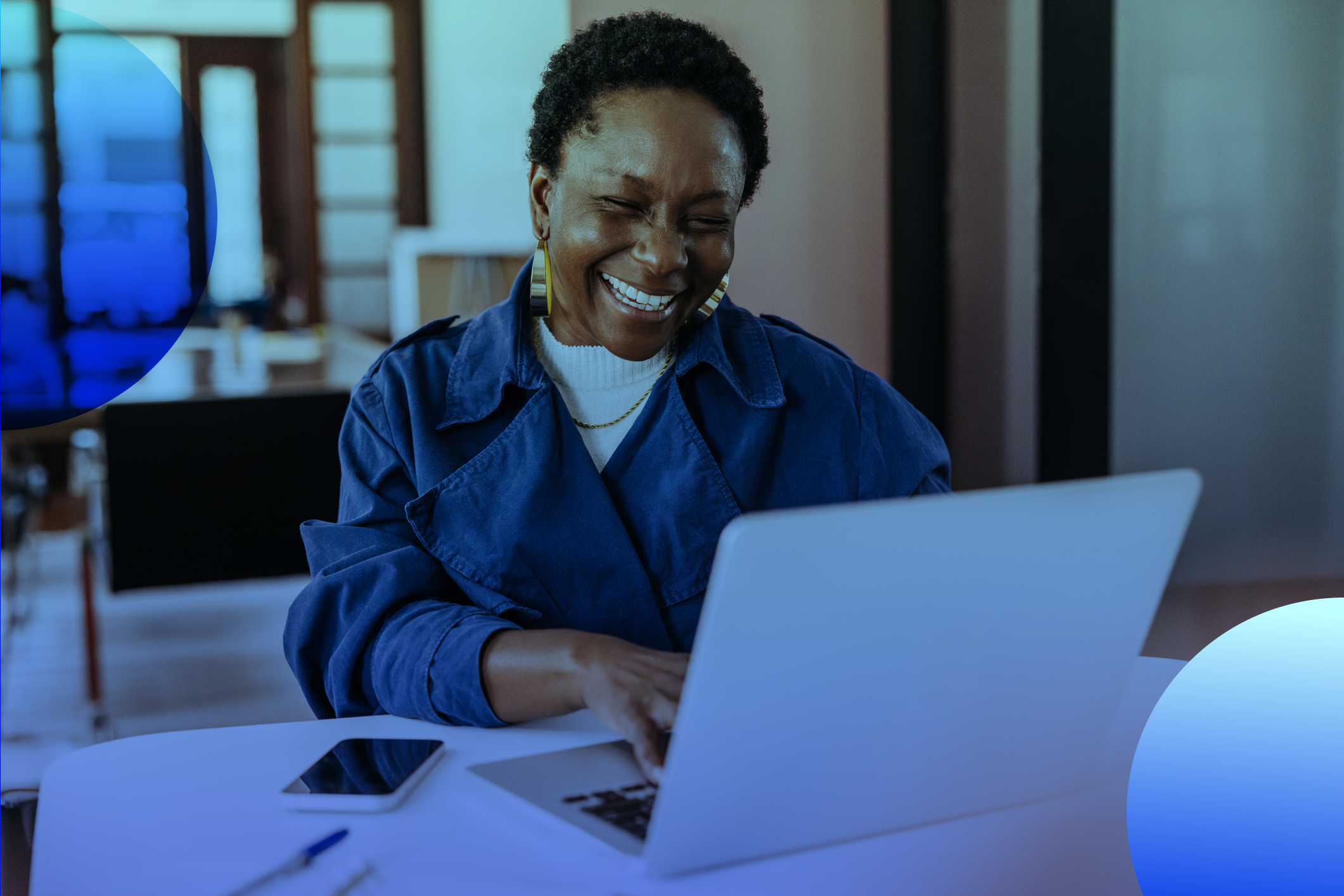 mulher negra vestida de camisa azul, sorrindo e usando um laptop sobre a mesa