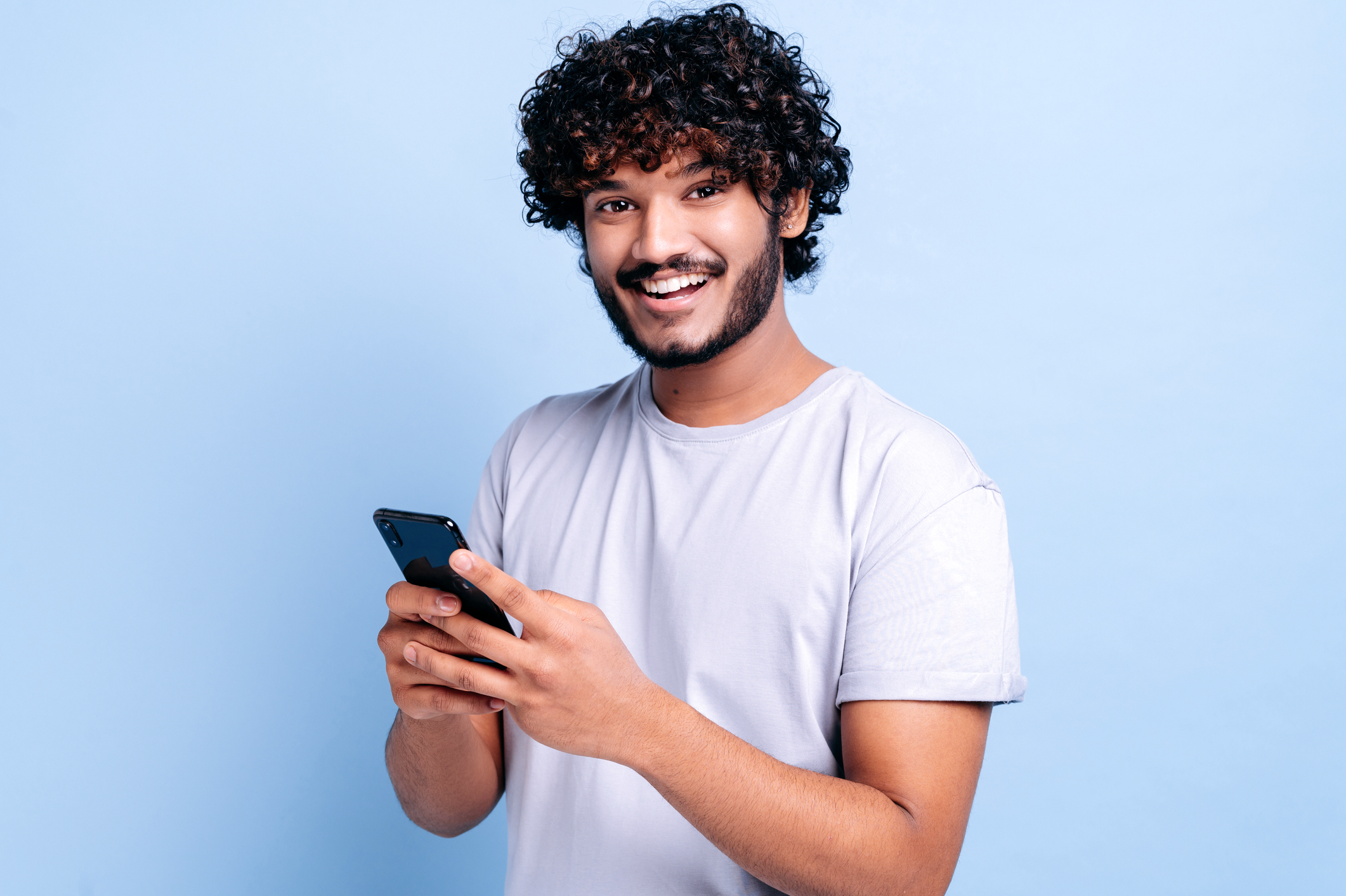 homem de jovem de cabelo cacheado e barba sorrindo com um smartphone nas mãos