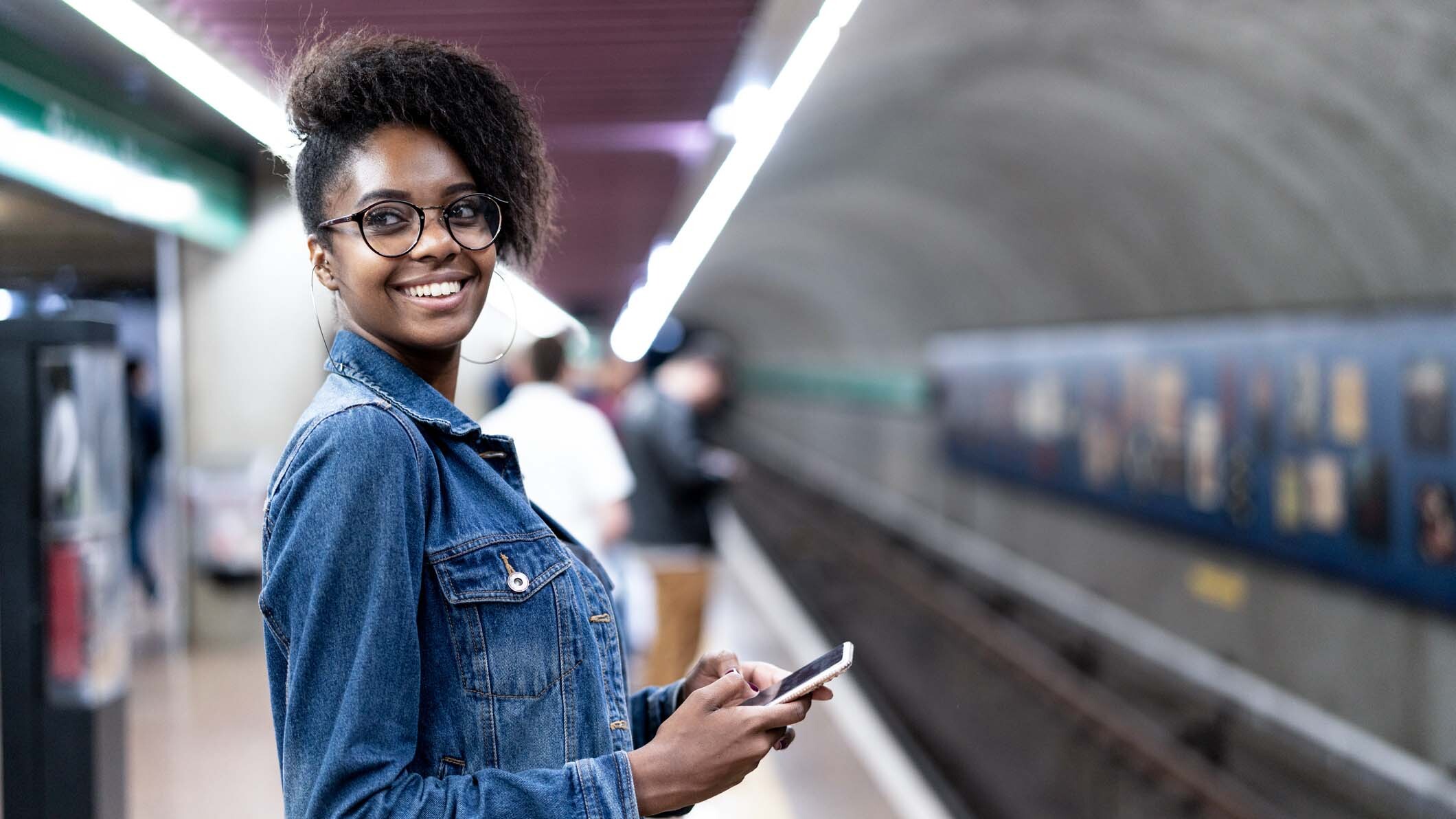 Mulher negra sorrindo em uma estação de metrô segurando um celular